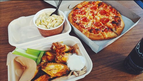 A takeout meal featuring pizza with pepperoni, chicken wings with celery, and a cup of pasta on a wooden table.