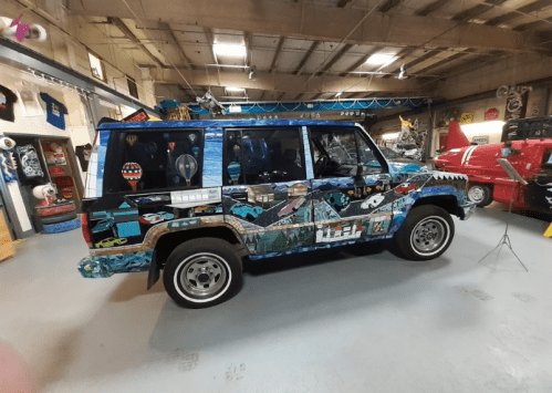 A colorful, mural-covered SUV displayed in a garage filled with vintage cars and memorabilia.