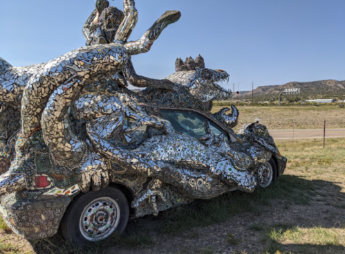 A mosaic-covered car adorned with sculptures of frogs and other creatures, set against a clear blue sky.