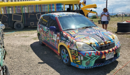 A colorful, artfully painted car with vibrant patterns parked on gravel, with a yellow bus in the background.