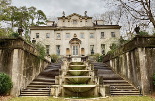 A grand, historic mansion with a staircase and fountain, surrounded by trees and greenery.