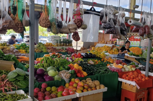A vibrant market stall filled with a variety of fresh fruits and vegetables, with vendors and shoppers in the background.