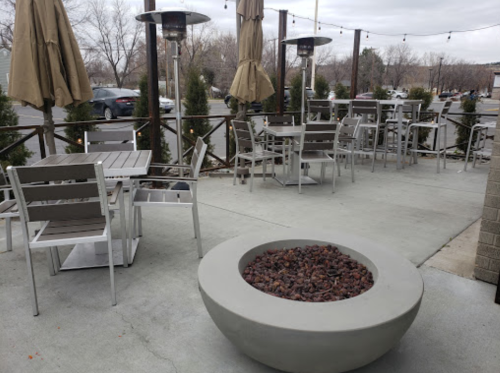 Outdoor seating area with metal tables and chairs, a fire pit, and patio umbrellas under a cloudy sky.