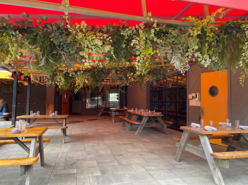 Outdoor dining area with wooden tables, greenery overhead, and a vibrant red canopy. Cozy and inviting atmosphere.
