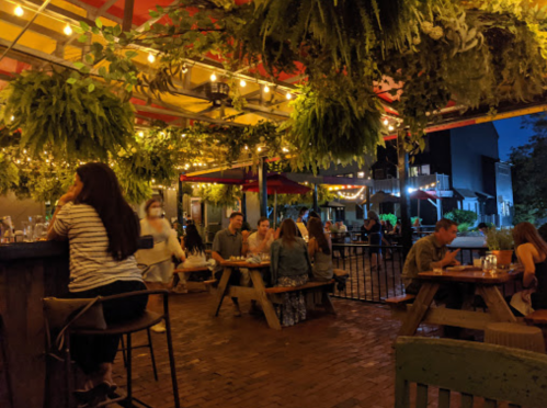 A lively outdoor dining area with string lights and hanging plants, featuring groups of people enjoying their meals at night.