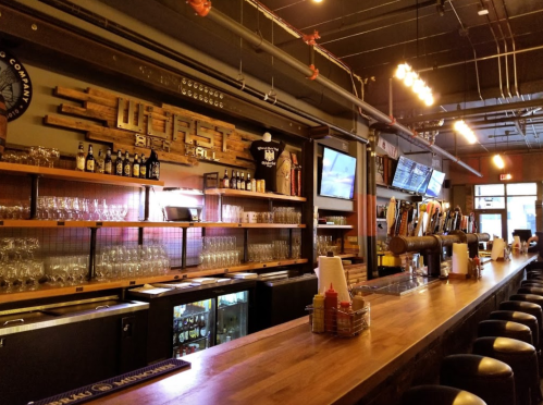 A cozy bar interior featuring a wooden counter, shelves of drinks, and a variety of beer taps under warm lighting.
