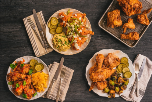 A spread of fried chicken, sides, and pickles on plates, with utensils and napkins on a dark wooden table.