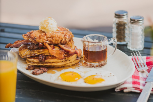 A plate of pancakes topped with fried chicken, bacon, and a butter pat, served with syrup and two sunny-side-up eggs.