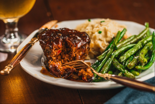 A plate featuring meatloaf with glaze, mashed potatoes, and green beans, alongside a glass of beer.