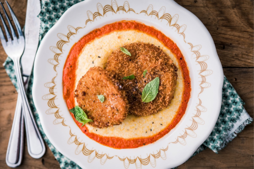 A plate with two crispy fried patties on a bed of creamy sauce, garnished with fresh herbs. Fork and knife beside it.