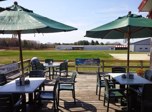 Outdoor dining area with green umbrellas overlooking a grassy field and a small airport in the background.