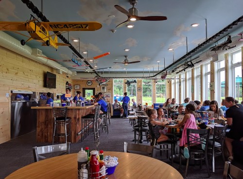 A lively restaurant interior with a bar, diners at tables, and model airplanes hanging from the ceiling.