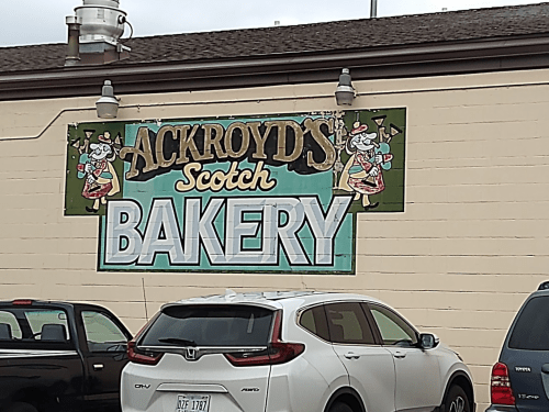 Sign for "Ackroyd's Scotch Bakery" featuring colorful graphics, with parked cars in front.