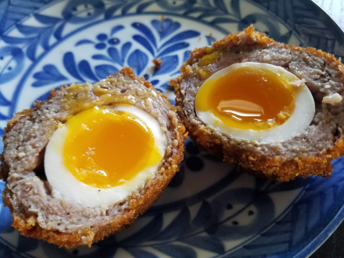 Two halves of a scotch egg on a decorative blue plate, showcasing a golden runny yolk surrounded by sausage meat.