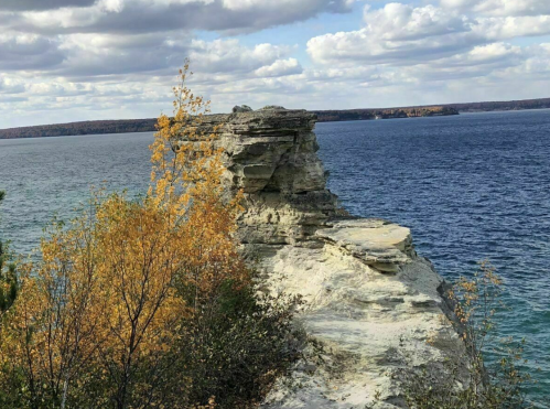 A rocky cliff overlooking a calm lake, surrounded by trees with autumn foliage under a cloudy sky.