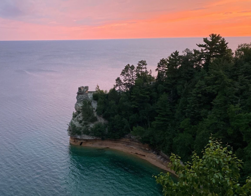 Sunset over a tranquil lake, highlighting a rocky cliff surrounded by lush trees and calm waters.