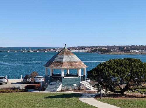 A gazebo by the water with a clear blue sky and distant shoreline, surrounded by green grass and a tree.
