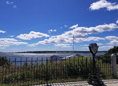 A scenic view of a waterfront with boats, under a bright blue sky filled with clouds, framed by a black fence.