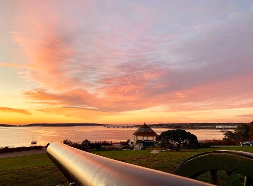 A scenic sunset over a calm waterway, with a gazebo and a cannon in the foreground, and colorful clouds in the sky.