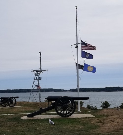 A cannon on a grassy area with flags at half-mast, overlooking a calm body of water and a distant shoreline.