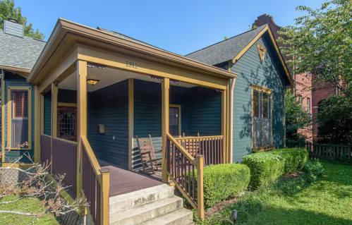 A blue house with a porch, featuring wooden railings and rocking chairs, surrounded by greenery.