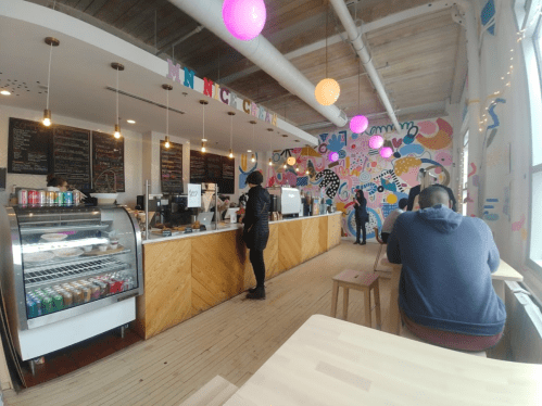 A colorful café interior with a counter, customers, and vibrant wall art. Bright pendant lights hang from the ceiling.