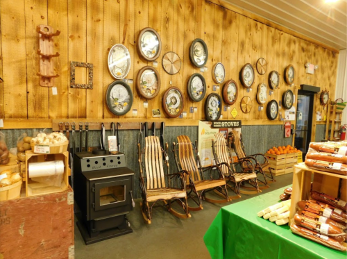 A rustic interior featuring wooden walls, decorative plates, rocking chairs, a stove, and a green table with products.