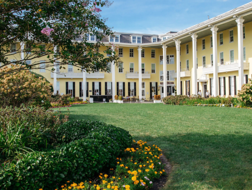A sunny courtyard with vibrant flowers, surrounded by a yellow building with white columns and outdoor seating.