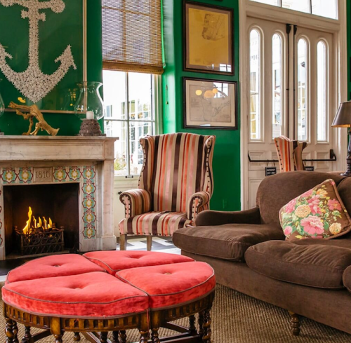 Cozy living room with green walls, striped chairs, a fireplace, and a pink ottoman in front of a large window.