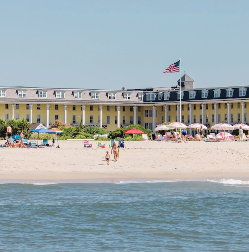 A beach scene with people walking on the sand, colorful umbrellas, and a large yellow building in the background.