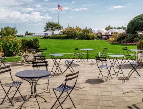 Outdoor patio with empty metal tables and chairs, lush green lawn, and an American flag in the background.