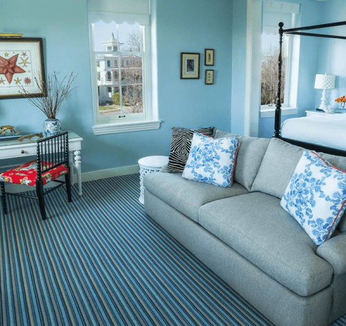 Cozy room with blue walls, striped carpet, a gray sofa, and a desk by a window, featuring decorative pillows and plants.