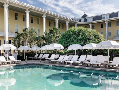 A serene pool area with lounge chairs and umbrellas, surrounded by greenery and a historic yellow building.