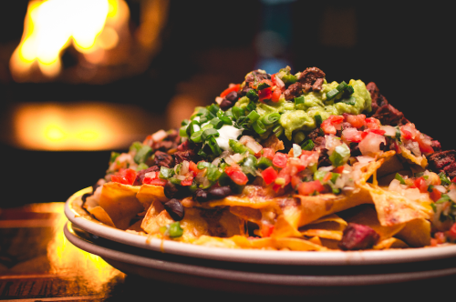 A plate of nachos topped with guacamole, diced tomatoes, green onions, and black beans, with a warm background.