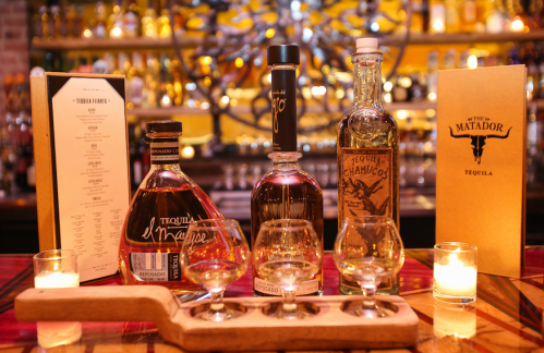A wooden flight with three tequila glasses, bottles of tequila, and a menu on a bar with soft candlelight.