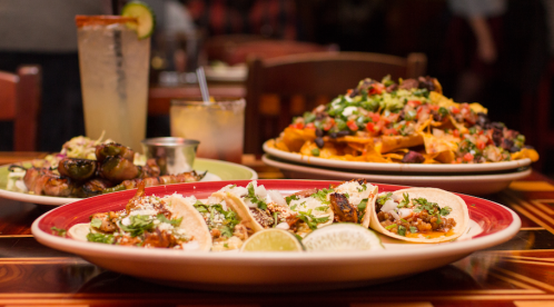 A plate of tacos with lime, alongside nachos and a drink, set on a wooden table in a restaurant.