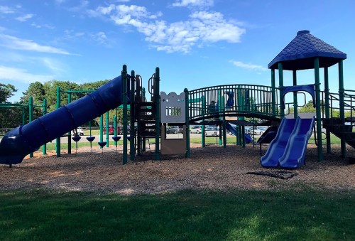 A colorful playground with slides, a bridge, and climbing structures, set on a grassy area under a blue sky.