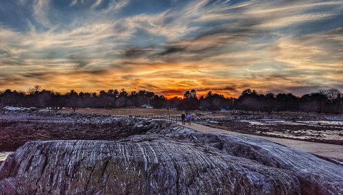 A rocky shoreline at sunset, with vibrant clouds and two people walking along a path in the distance.
