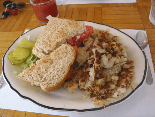 A plate with a sandwich containing lettuce and tomato, alongside crispy hash browns and pickles.
