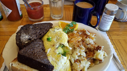 A plate of breakfast featuring an omelet, hash browns, toast, orange slice, coffee, and condiments on a wooden table.