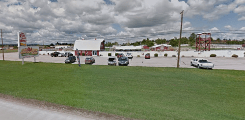 A roadside view of a red building with a sign, surrounded by parked cars and a grassy area under a cloudy sky.