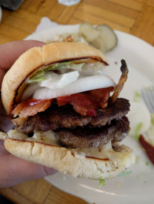 A hand holding a stacked burger with two patties, bacon, lettuce, and onions, served on a toasted bun.