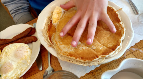 A hand rests on a large pancake on a plate, with bacon and eggs on the side.