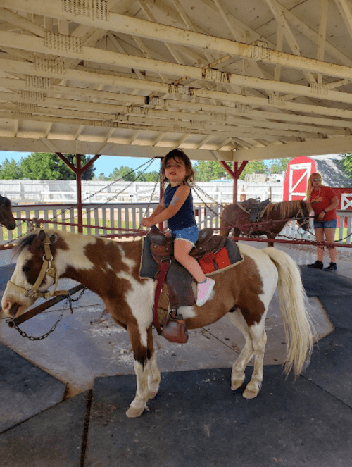 A young girl smiles while riding a pony on a carousel, with another person standing nearby in a barn setting.