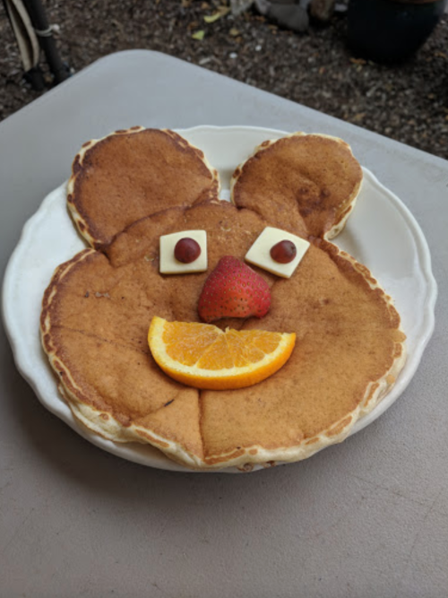 A plate of pancakes arranged to look like a bear face, with fruit and chocolate for eyes and a smile.
