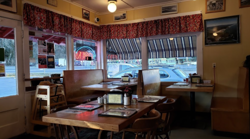Cozy diner interior with wooden tables, chairs, and large windows, featuring red and white patterned curtains.