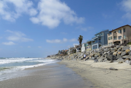 A sandy beach with waves, rocky shore, and colorful houses along the coastline under a blue sky with clouds.