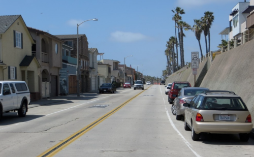 A coastal street lined with houses and parked cars, with palm trees and a clear blue sky in the background.