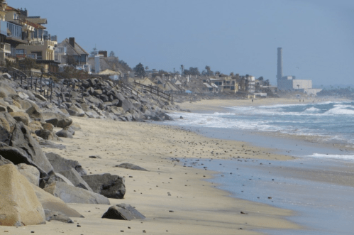 A sandy beach lined with rocks and houses, with waves gently crashing and a distant industrial structure.