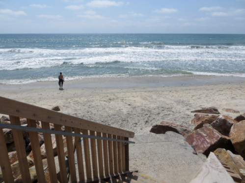 A person walks along a sandy beach with waves crashing, viewed from a staircase leading down to the shore.
